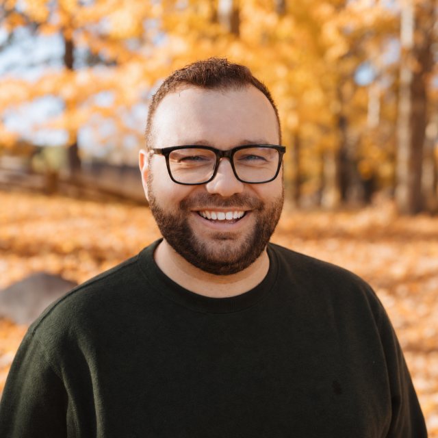 Jarid Hill, smiling, in glasses and a dark sweater with autumn trees behind him